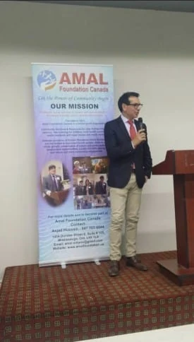 Asjad Hussain speaking beside Amal Foundation Canada materials